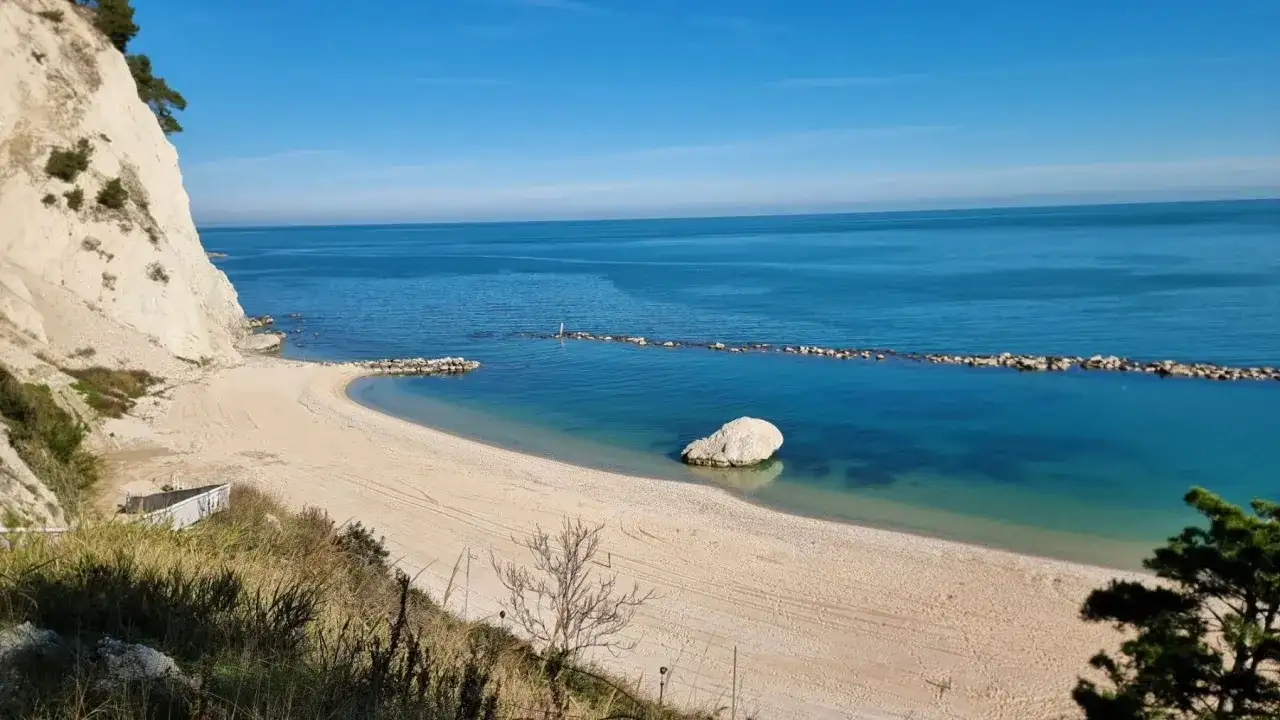 Spiaggia dorata e mare cristallino sulla Riviera del Conero, con una scogliera bianca a picco sull'acqua e un grande masso solitario.