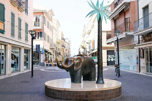Scultura di elefante e palma sul centro passeggiata San Benedetto del Tronto, con negozi e palazzi colorati sullo sfondo.