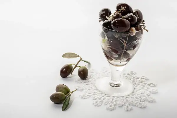 Un bicchiere pieno di olive nere, con rametti e foglie, accanto a olive fresche su un pizzo. Le olive delle Marche, un sapore autentico.