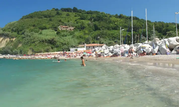 Spiaggia affollata con persone che nuotano e prendono il sole, sullo sfondo una collina verdeggiante. Gabicce Mare, dove si trova questo angolo di paradiso, è una perla dell'Adriatico.
